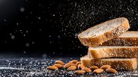 Fresh sliced white bread stacked with scattered flour dust and raw almonds on dark background, dramatic food photography with copy space.