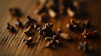 Aromatic cloves scattered on wooden surface in warm brown tones with selective focus and dramatic lighting, creating moody culinary atmosphere.