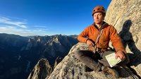 Male mountaineer in orange jacket and helmet reading map on rocky summit with dramatic mountain landscape and blue sky background for outdoor adventure content.