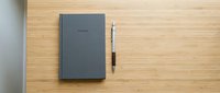 Closed dark gray notebook with pen on wooden desk surface, flat lay composition for business and office design projects.