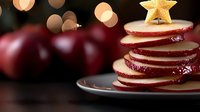 Festive sliced red and white apple stack arranged as Christmas tree topped with golden star decoration on dark plate with bokeh lights background.
