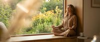 Young woman in beige loungewear sitting peacefully by large window with morning sunlight streaming in, perfect for wellness and mindfulness content.