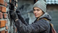 Young Caucasian woman in winter clothing and protective gloves working on brick wall renovation, smiling while doing construction work outdoors in cold weather.