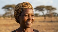 Smiling African woman wearing traditional headwrap in savanna landscape with acacia trees, representing cultural heritage and rural community life.