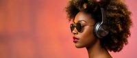 Young African American woman with curly afro hair wearing sunglasses and headphones against vibrant coral pink background for music and lifestyle content.