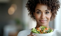 Young African American woman with curly hair smiling while holding fresh vegetable salad bowl, representing healthy eating lifestyle and wellness.