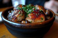 Glazed chicken thighs in rustic ceramic bowl garnished with fresh herbs and chickpeas, served in traditional style with caramelized sauce and vibrant greens.