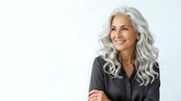 Mature woman with long silver wavy hair wearing dark button-up shirt, smiling confidently against white background. Suitable for business, healthcare, corporate websites and professional portraits.
