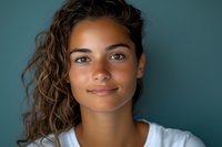 Young mixed race woman with curly hair and natural makeup smiling confidently against teal background, wearing white top. Soft studio lighting creates gentle shadows.