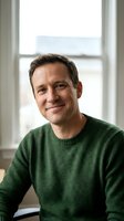 Confident middle-aged Caucasian man in green sweater smiling warmly by window in bright indoor setting for business and lifestyle content.