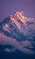 Majestic snow-capped mountain peak illuminated by golden sunrise light against purple sky, rising above misty clouds in dramatic alpine landscape.