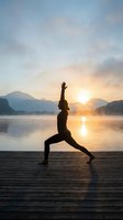Woman practicing yoga warrior pose on wooden dock at sunrise over mountain lake with misty morning atmosphere and golden sunlight reflection.