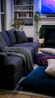 Cozy living room interior with dark blue upholstered sofa, decorative pillows, knit throw blanket, potted plants, wall shelving, fireplace, and glowing screen for home decor and interior design projects.