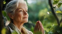 Senior woman with silver hair meditating in lush green garden, hands in prayer position, peaceful expression, soft natural lighting creates serene atmosphere.