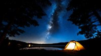 Camping tent glowing under starry night sky with Milky Way galaxy over mountain lake. Outdoor adventure and wilderness exploration concept.