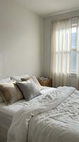Minimalist bedroom interior with unmade bed, neutral linen bedding, soft natural lighting through sheer curtains creating cozy morning atmosphere.