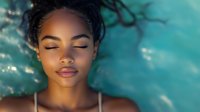 Young African American woman with closed eyes and serene expression floating in turquoise water. Natural makeup, glowing skin and peaceful demeanor create tranquil spa atmosphere.