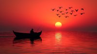 Silhouette of lone fisherman in wooden boat against dramatic red sunset sky with flying birds and sun reflection on calm water surface.