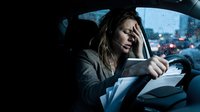 Stressed businesswoman driver holding head while sitting in car during rainy evening traffic jam with city lights blurred in background.