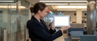 Businesswoman with dark hair and fair complexion wearing black blazer holding laptop with blank screen in modern office environment, suitable for corporate, technology, and business presentations.