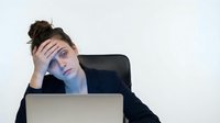 Young stressed businesswoman with hand on forehead showing exhaustion and burnout while working on laptop in modern office environment.