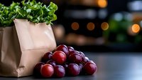 Fresh red grapes clustered on dark surface with kraft paper grocery bag and green lettuce leaves, soft bokeh lights in background creating cozy atmosphere.