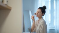 Young woman applying lipstick in modern bathroom mirror during morning beauty routine. Natural lighting creates soft atmosphere for skincare and makeup.