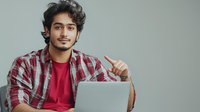 Young South Asian man in plaid shirt and red t-shirt showing thumbs up while working on laptop, expressing positive attitude and satisfaction. Professional workplace setting.