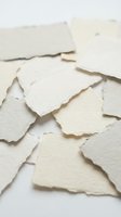 Torn pieces of cream colored handmade paper with rough deckled edges scattered on white surface for crafting and artistic projects.