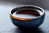 Traditional ceramic bowl with dark blue glaze exterior and amber liquid inside, photographed on black slate surface with dramatic side lighting creating reflections.