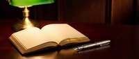 Open book with fountain pen on wooden desk under green banker lamp creating warm study atmosphere for reading and writing.
