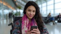 Young Hispanic woman in wheelchair using smartphone at airport terminal, wearing colorful floral jacket and pink scarf, expressing joy and independence.