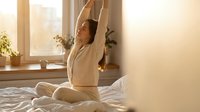 Young woman stretching arms up while sitting on bed in bright morning sunlight streaming through window creating peaceful wellness moment.
