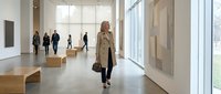 Woman in beige coat walking through modern art gallery with wooden benches and large windows, contemporary museum interior with visitors exploring exhibitions.