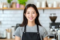 Young Asian female barista wearing gray apron and t-shirt smiling at camera in modern coffee shop interior with coffee equipment and shelving in background.