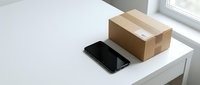 Smartphone and cardboard box on white desk near window, suitable for e-commerce, delivery, and online shopping concepts.