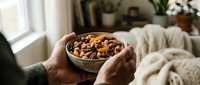 Elderly hands holding wooden bowl filled with mixed nuts including walnuts, almonds and hazelnuts in cozy home setting with soft natural lighting.