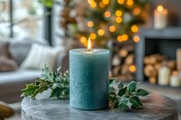 Burning teal pillar candle with holly leaves on concrete surface against blurred Christmas tree lights creating cozy festive atmosphere for winter holidays.