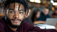 Young African American male student with dreadlocks and glasses smiling confidently in university library or study space, wearing burgundy sweater. Blurred background.