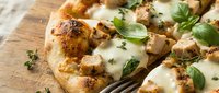 Baked flatbread topped with melted cheese, roasted chicken pieces, and fresh basil garnish on wooden surface for food photography and culinary content.