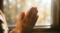 Woman hands pressed together in prayer position with warm sunlight streaming through window creating peaceful spiritual moment for meditation and mindfulness practice.