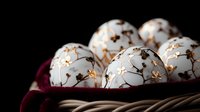 White porcelain Easter eggs with delicate gold floral pattern arranged in woven basket with burgundy fabric, dramatic lighting against dark background.