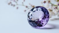 Round cut purple amethyst gemstone with brilliant facets casting soft shadows, displayed on white surface with delicate floral elements in background.