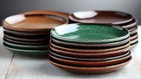 Stacks of handmade ceramic plates and bowls in earthy brown and green tones on rustic wooden surface, showcasing artisanal pottery craftsmanship and tableware design.