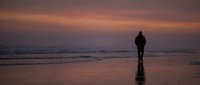 Silhouetted figure standing alone on wet beach sand during golden hour sunset with calm ocean waves and dramatic sky backdrop.