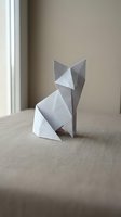White paper origami cat sitting on beige fabric surface near window with soft natural lighting for craft and hobby projects.