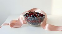 Young woman's hands holding glass bowl filled with fresh mixed berries including blueberries and raspberries, decorated with pink satin ribbon, isolated on white background for food and wellness design projects.