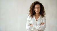 Young mixed-race businesswoman with curly hair smiling confidently with crossed arms in white shirt against neutral background for corporate use.