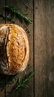 Freshly baked artisan sourdough bread loaf with golden crust on rustic wooden table with rosemary herbs for bakery, restaurant menu and cooking concepts.
