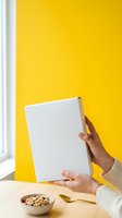 Cropped view of a person with medium skin tone holding a blank white board against a vibrant yellow wall, with a bowl of cereal and gold spoon on wooden table for advertising and product mockup use.